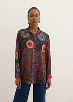A woman modeling a long-sleeve button-up shirt with a vibrant, multicolored circular mandala pattern in red, orange, blue, and green on a dark base, paired with dark blue jeans against a plain light beige background.