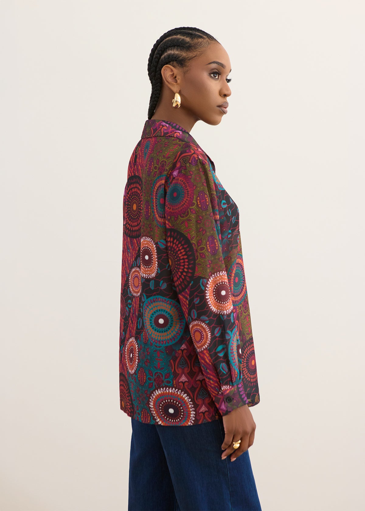 A woman shown in side profile wearing a long-sleeve button-up shirt with a vibrant, multicolored circular African wax print, paired with dark blue pants, against a plain off-white background.