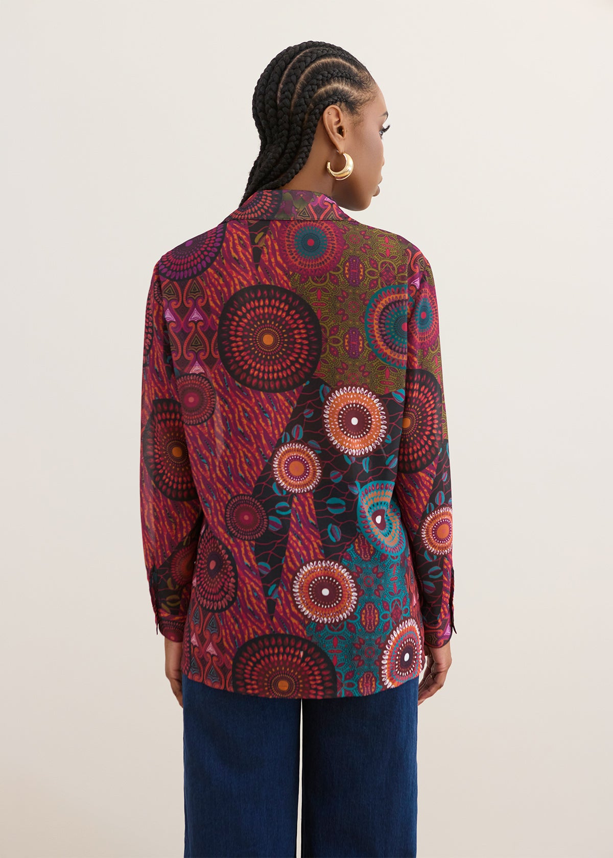 A woman shown from the back wearing a long-sleeve button-up shirt with a vibrant, multicolored circular mandala pattern in red, green, blue, and black, paired with dark blue jeans against a plain light beige background.