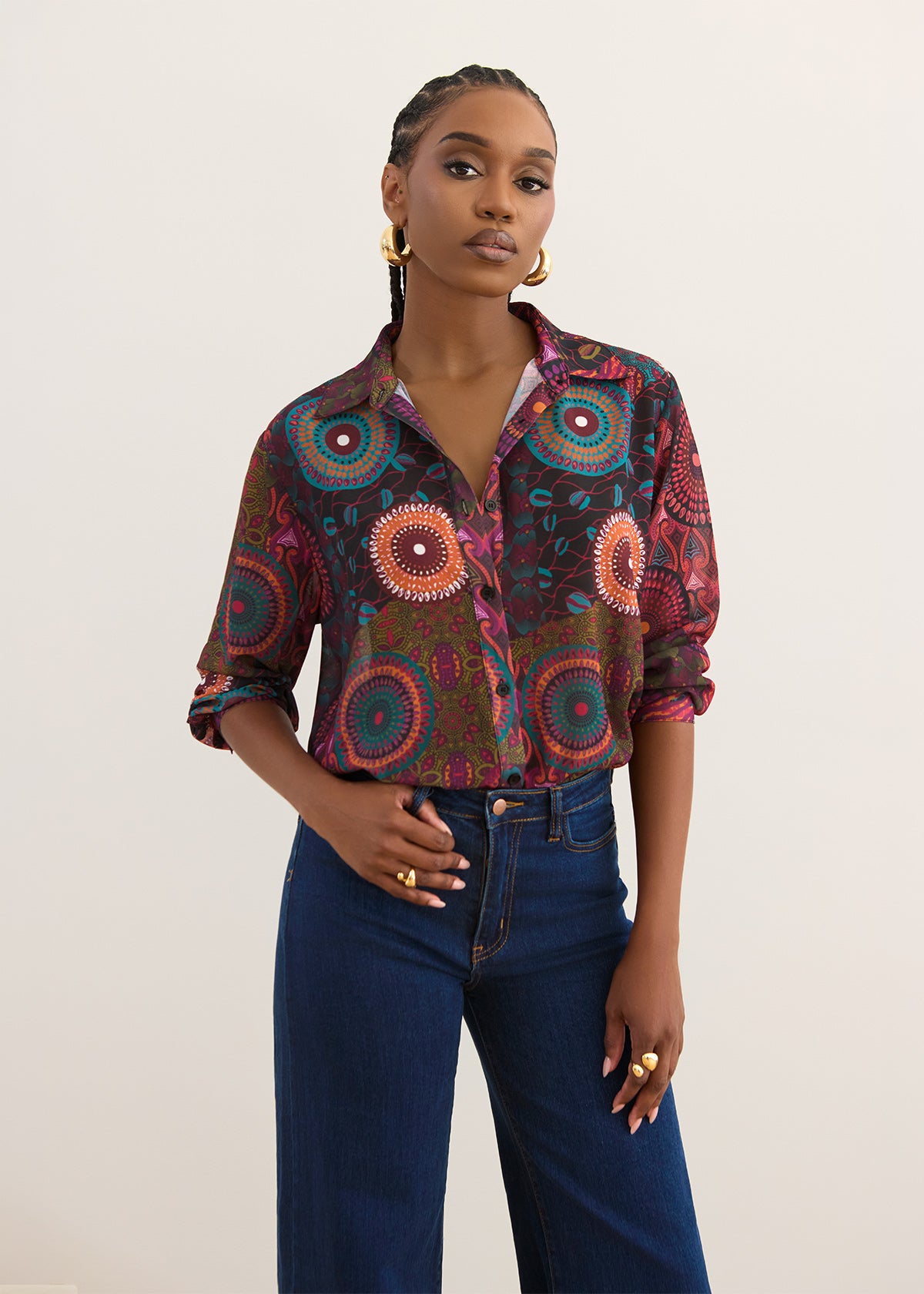 A woman modeling a long-sleeve button-up shirt with a colorful circular mandala pattern in red, orange, teal, and brown, paired with dark blue jeans against a plain off-white background.