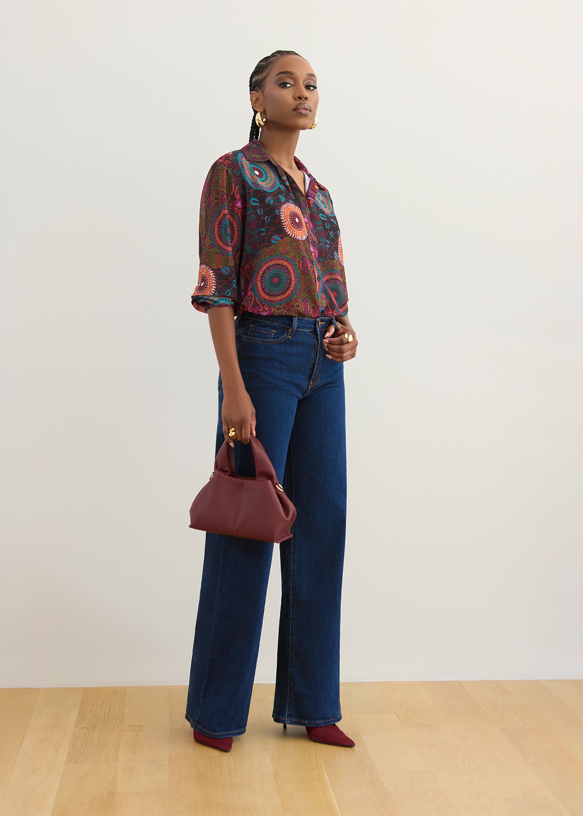 A model stands in a studio wearing a colorful circular-patterned button-up blouse, dark blue wide-leg jeans, burgundy pointed-toe heels, and holding a burgundy handbag.
