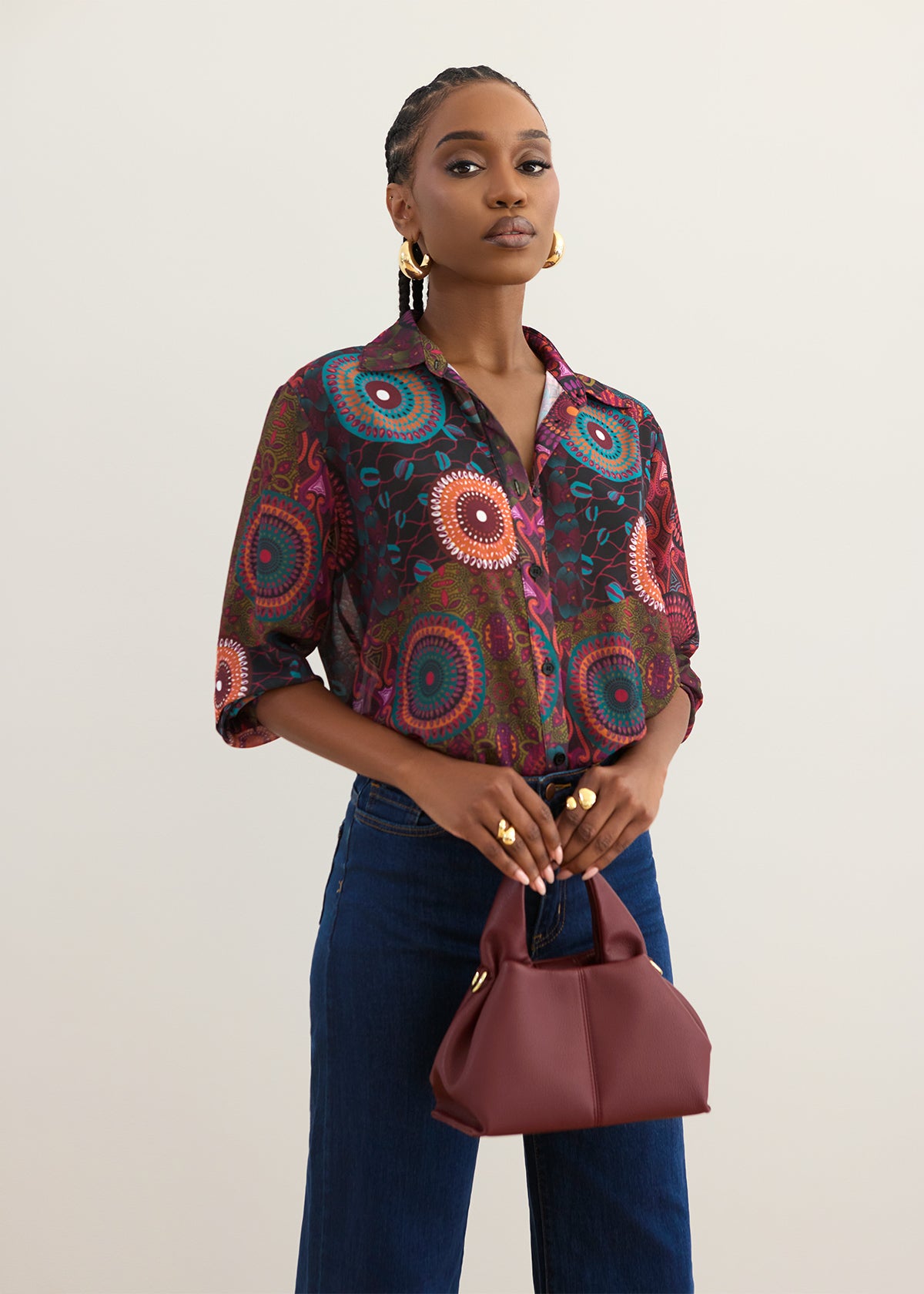 A woman modeling a long-sleeve button-up shirt with a vibrant multicolor circular mandala print, paired with dark blue jeans and holding a burgundy handbag against a plain light beige background.