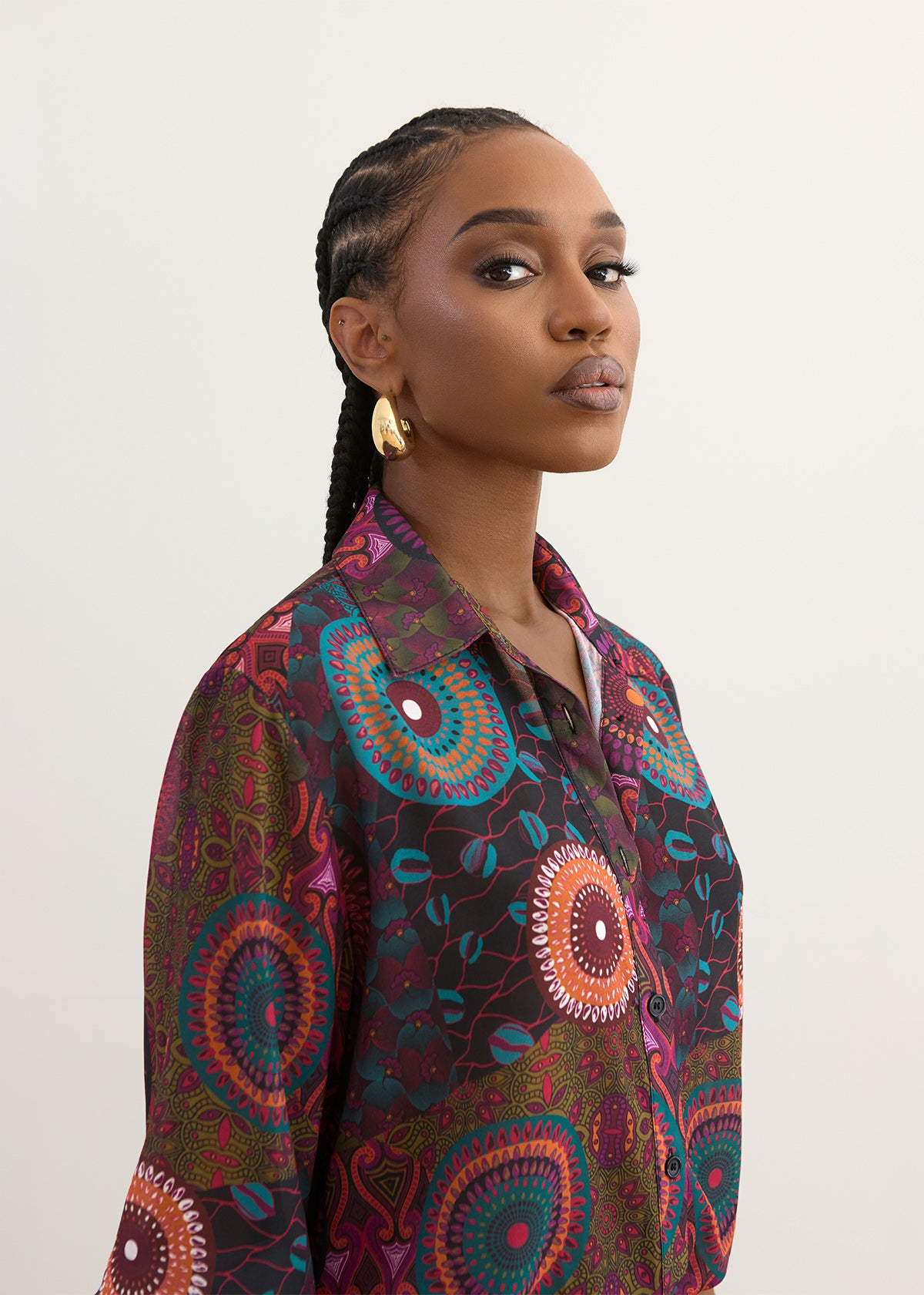 A woman wearing a long-sleeve button-up shirt with a vibrant, multicolored circular mandala pattern in shades of purple, teal, orange, and green, photographed against a plain off-white background.
